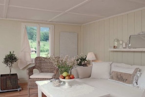 Une pièce avec un canapé blanc, un fauteuil en osier, une table sur laquelle se trouve un panier de fruits, une lampe et une vue sur la verdure à l’extérieur.
