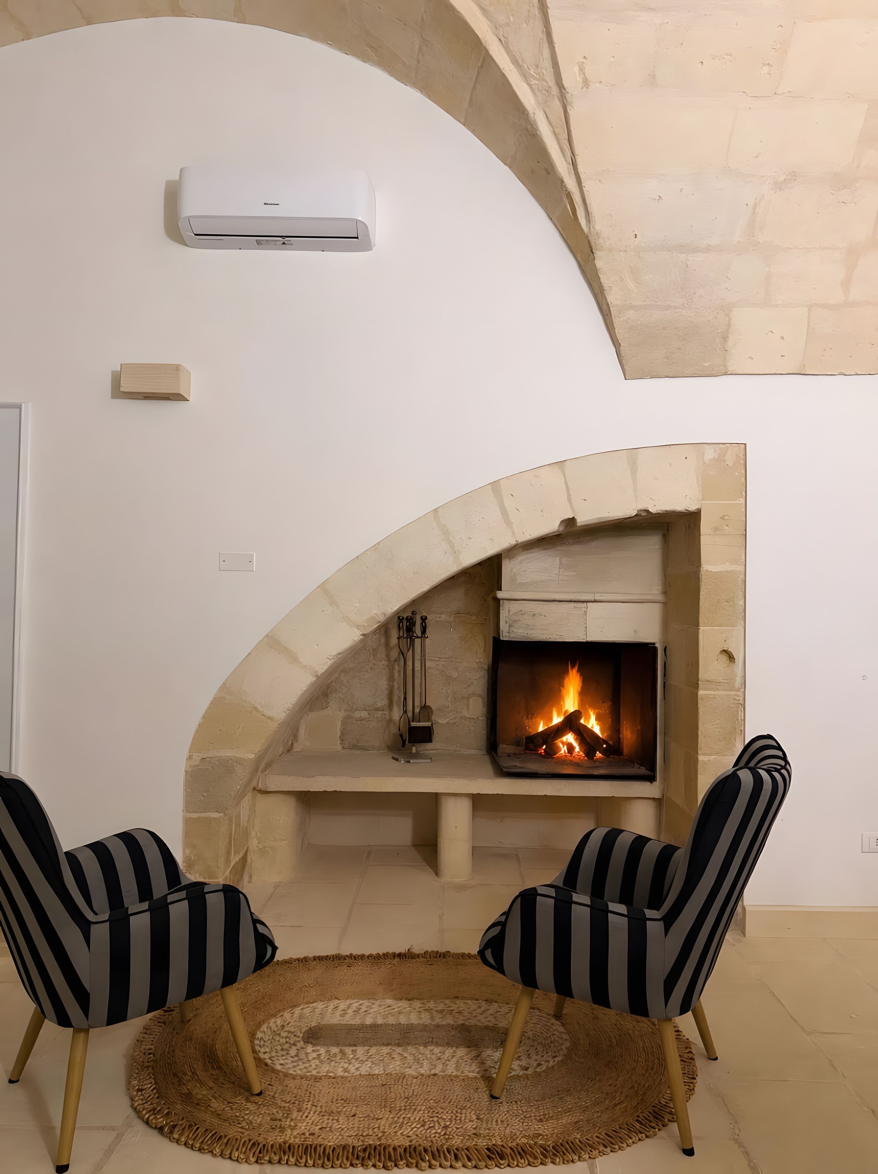 Une pièce avec une cheminée en pierre, deux fauteuils à rayures, un tapis tissé et un appareil de climatisation.