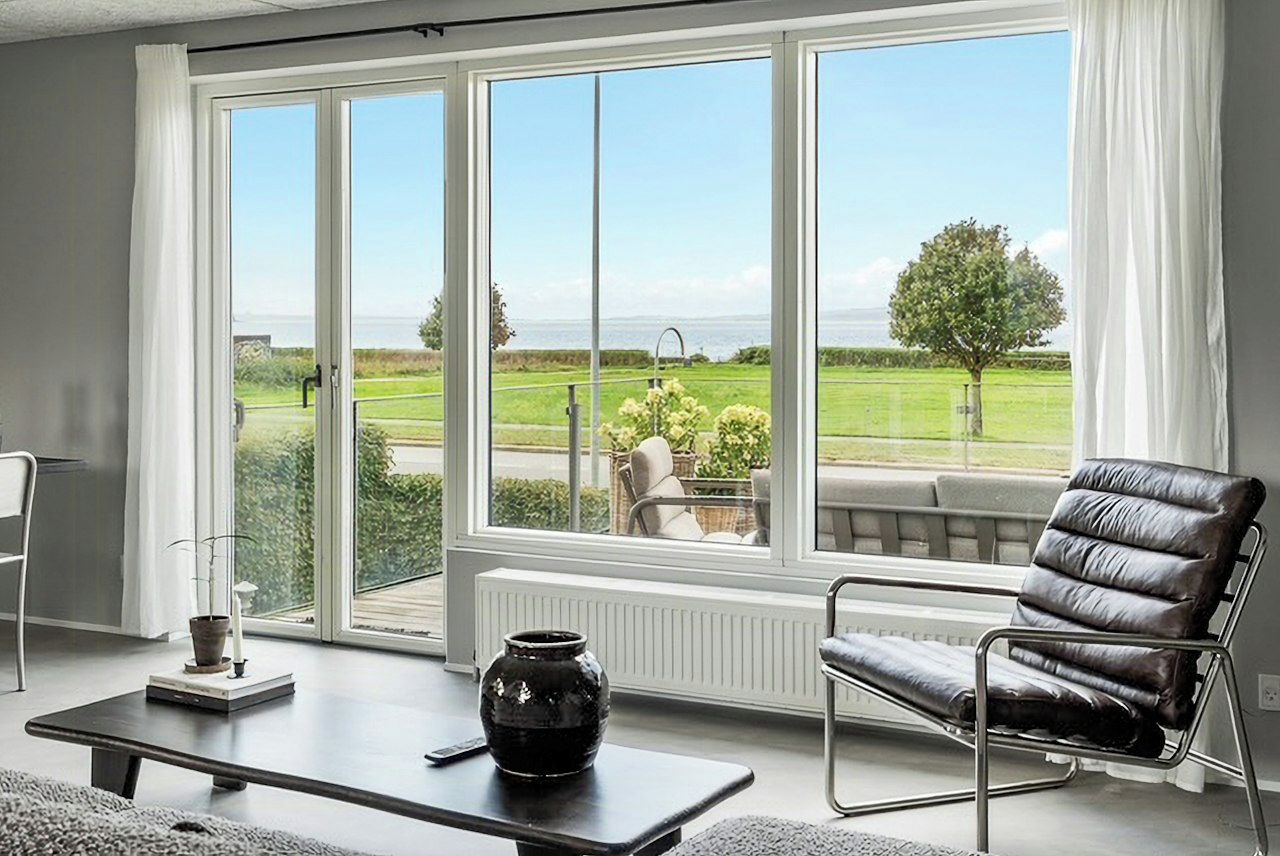 A room with a large window, a black leather chair, a small table with a vase, and a view of a green landscape.