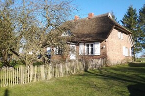 A traditional brick house with a thatched roof, surrounded by a wooden fence and trees. - Thatched house by the pond (Rubenow \/ Voddow)