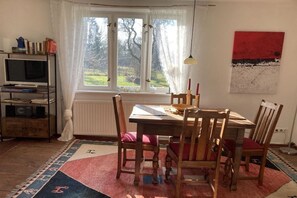 A dining area with a table, chairs, a radiator, a bookshelf, and a window with curtains. - Thatched house by the pond (Rubenow \/ Voddow)