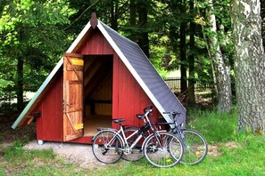 Eine kleine, rote Holzhütte mit geneigtem Dach, vor der zwei Fahrräder stehen, mit einem bewaldeten Hintergrund.