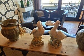 A room with a stone wall, a fireplace, a wooden table with bird sculptures, and a view of the outside through large windows. - Pug holiday pure holiday with south-facing balcony 009 (Burgtiefe-Südstrand)