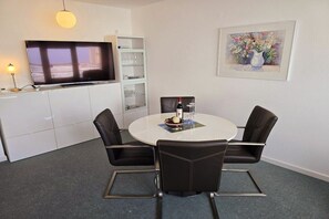Ein modernes Zimmer mit Fernseher, einem runden Esstisch mit vier Stühlen, einem weißen Schrank mit Glastüren und einem gerahmten Gemälde an der Wand.