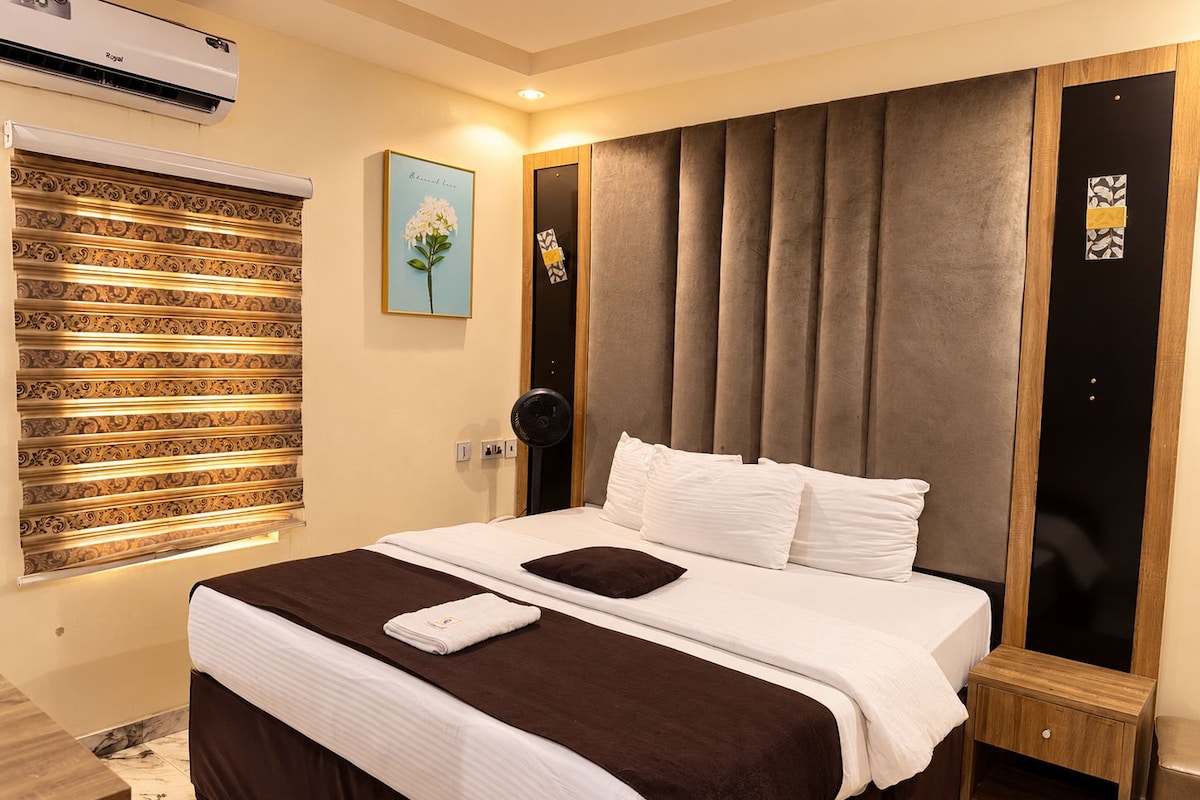 Une chambre d’hôtel comprenant un lit, une table de chevet, un appareil de climatisation, une fenêtre avec des stores et une œuvre d’art accrochée au mur.