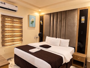 Une chambre d’hôtel comprenant un lit, une table de chevet, un appareil de climatisation, une fenêtre avec des stores et une œuvre d’art accrochée au mur.