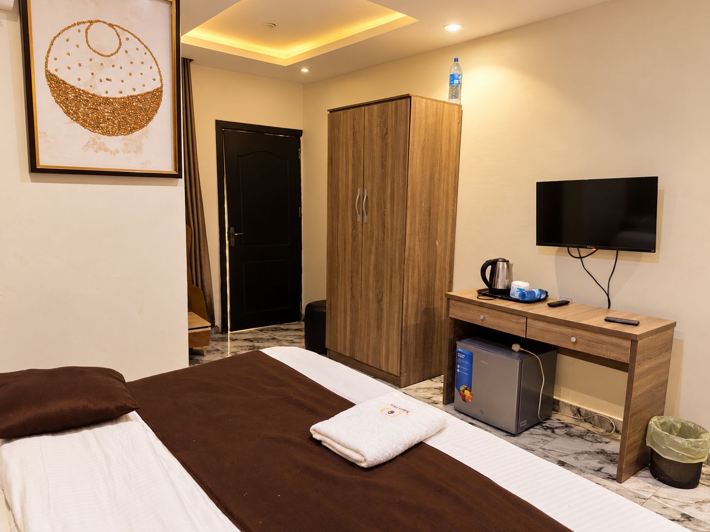 Habitación de hotel con cama, armario de madera, escritorio con televisor, hervidor de agua y botella.