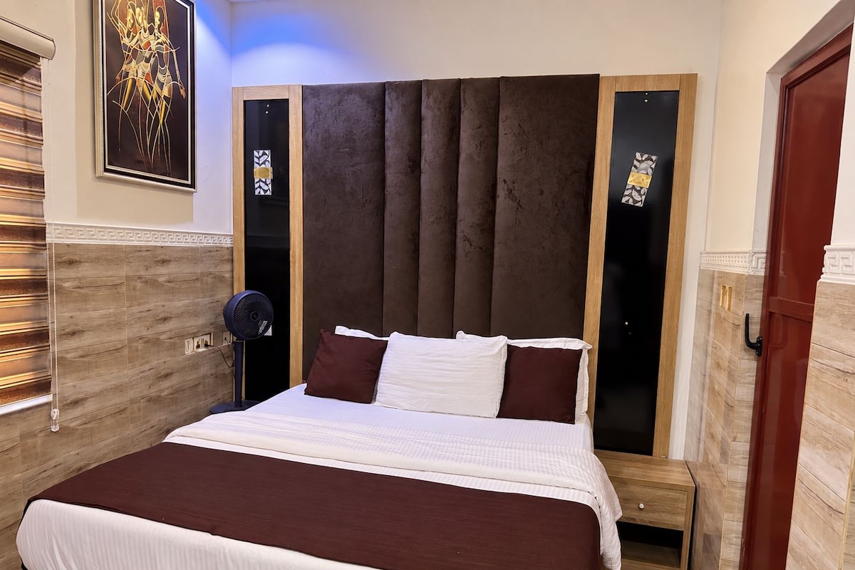 Une chambre d’hôtel avec un grand lit, une tête de lit en bois, un ventilateur et une porte dotée d’une poignée.