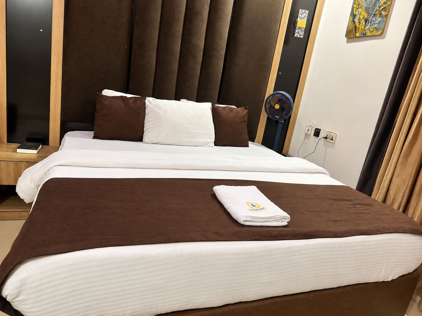 A hotel room with a bed, pillows, a fan, and a towel.