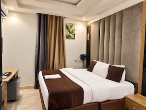 Une chambre d’hôtel avec un lit, un bureau, une chaise, un ventilateur et un tableau au mur.