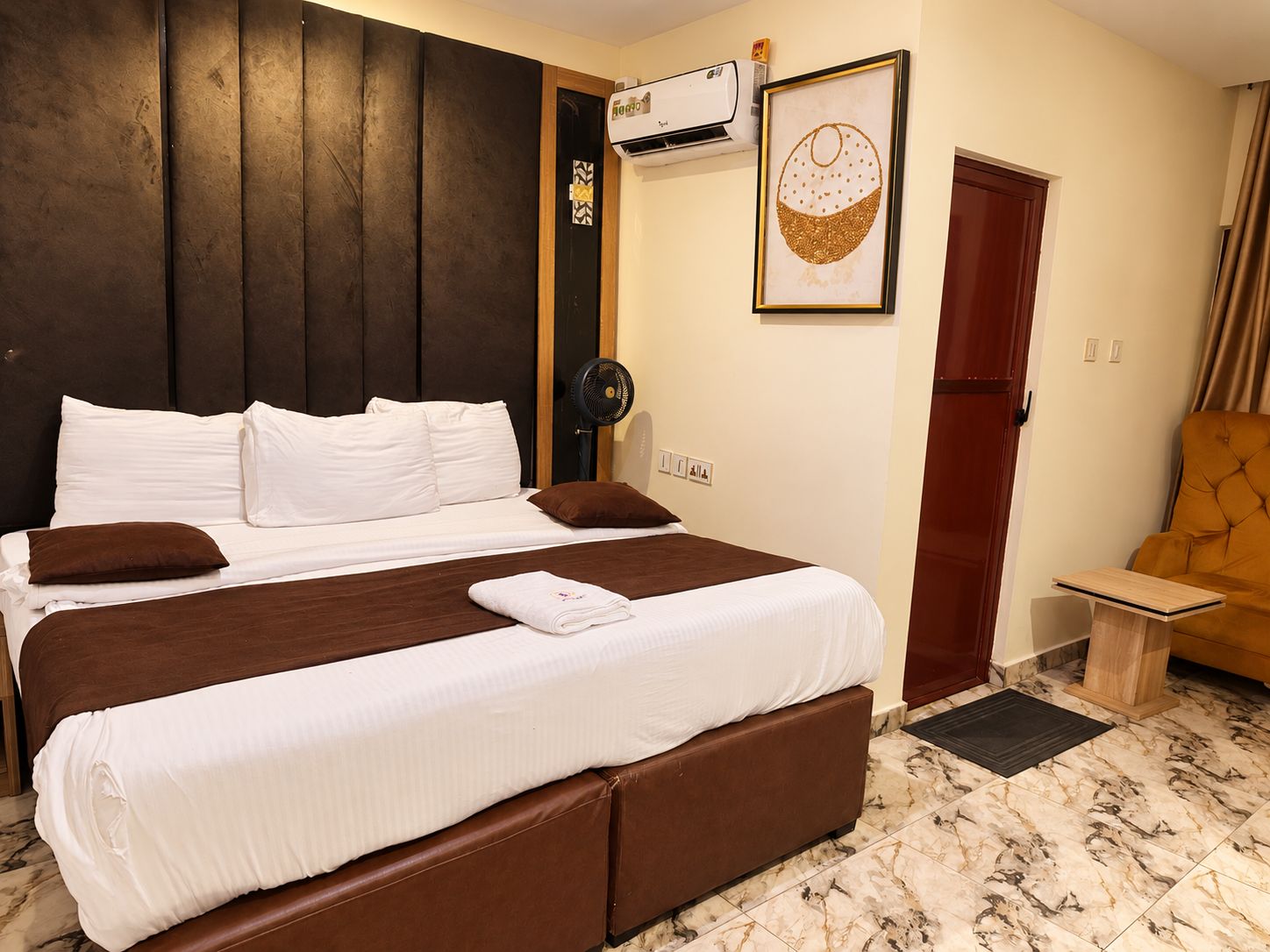 Una habitación de hotel con una cama grande, un ventilador junto a la cama, una puerta y una mesita.