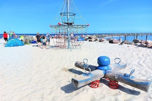 A sandy beach with a swing set, a rope swing, and a blue ball. - Holiday home in Damp with swimming pool (Damp)