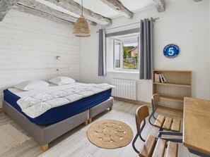 A bedroom with a bed, a wooden chair, a round woven rug, a bookshelf, and a window with curtains. - House for 8 with heated pool (La Bachellerie)
