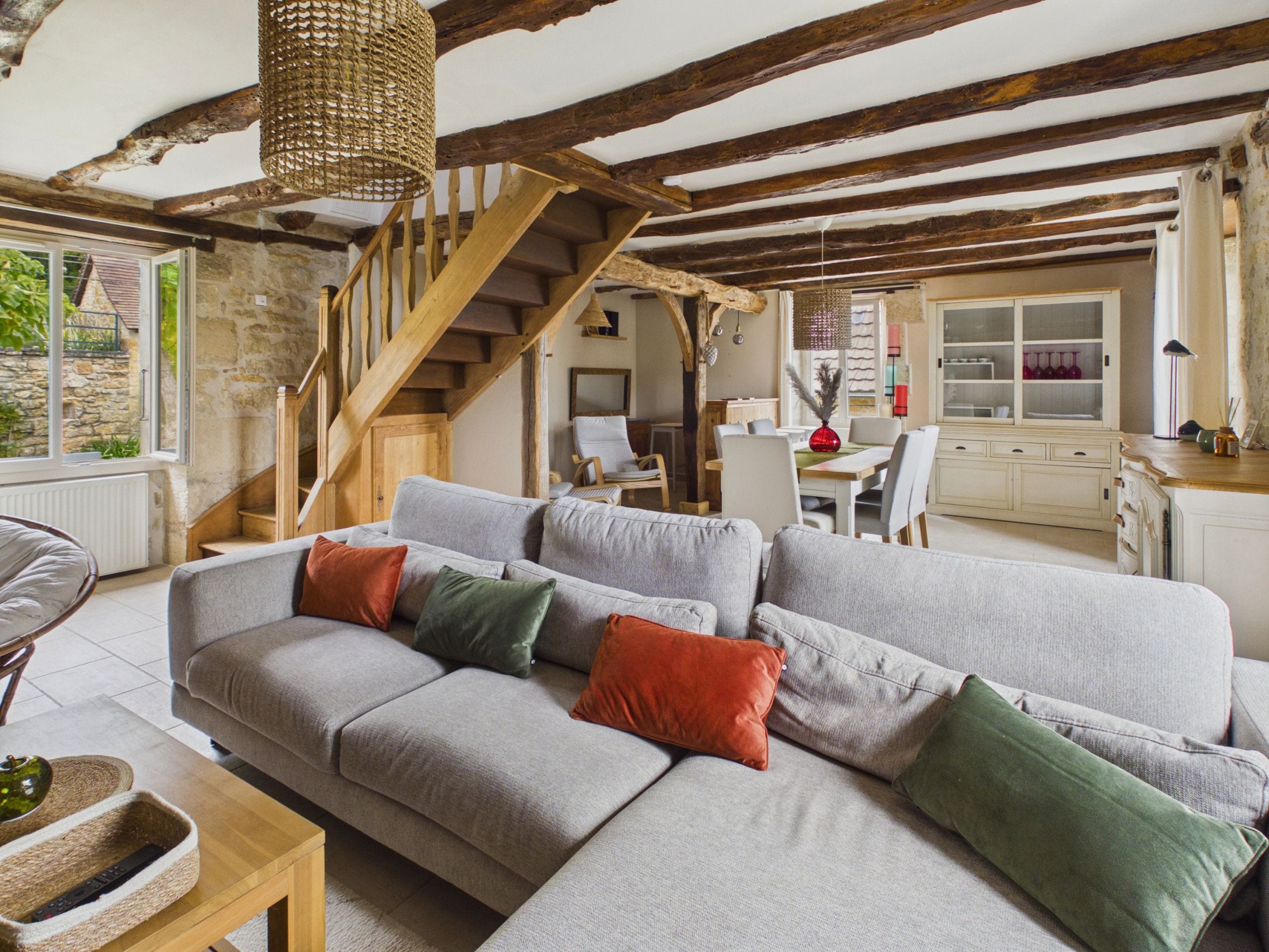 Een moderne woonkamer met een grijze hoekbank, houten balken in het plafond, een trap en een eethoek met witte meubels.
