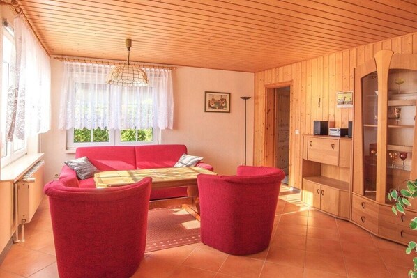 Un salon chaleureux avec un canapé rouge, deux fauteuils rouges, un meuble en bois et une fenêtre ornée de rideaux blancs.