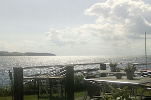 Une terrasse en bord de mer, avec des tables et des chaises, d’où l’on peut admirer un plan d’eau et la côte au loin.
