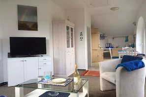 A living room with a television, a glass coffee table, and a dining area with a chair and table. - Holiday apartment with 2 bedrooms (Schönberger Strand)