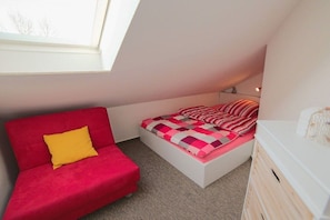 A small, compact room with a sloped ceiling, a single bed with a checkered bedspread, a red sofa with a yellow pillow, and a white dresser. - Holiday apartment with 2 bedrooms (Schönberger Strand)