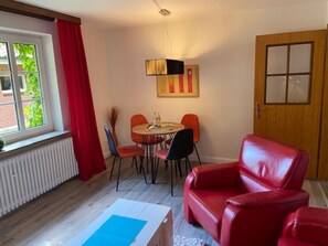 Ein Zimmer mit einem roten Ledersofa, einem Esstisch mit Stühlen, einem Heizkörper, einem Fenster mit roten Vorhängen und einer Tür mit Fensterteil.