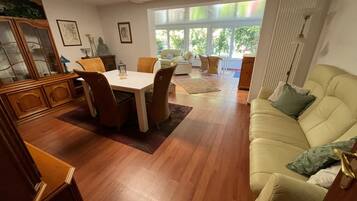 En rummelig opholdsstue med spisebord, stole, sofa og et stort vindue, der lukker naturligt lys ind.