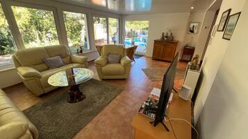 A modern living room with a sofa, armchair, and coffee table, a television on a stand, and large windows with a view of the outdoors.