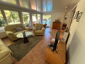 A modern living room with a sofa, armchair, and coffee table, a television on a stand, and large windows with a view of the outdoors.
