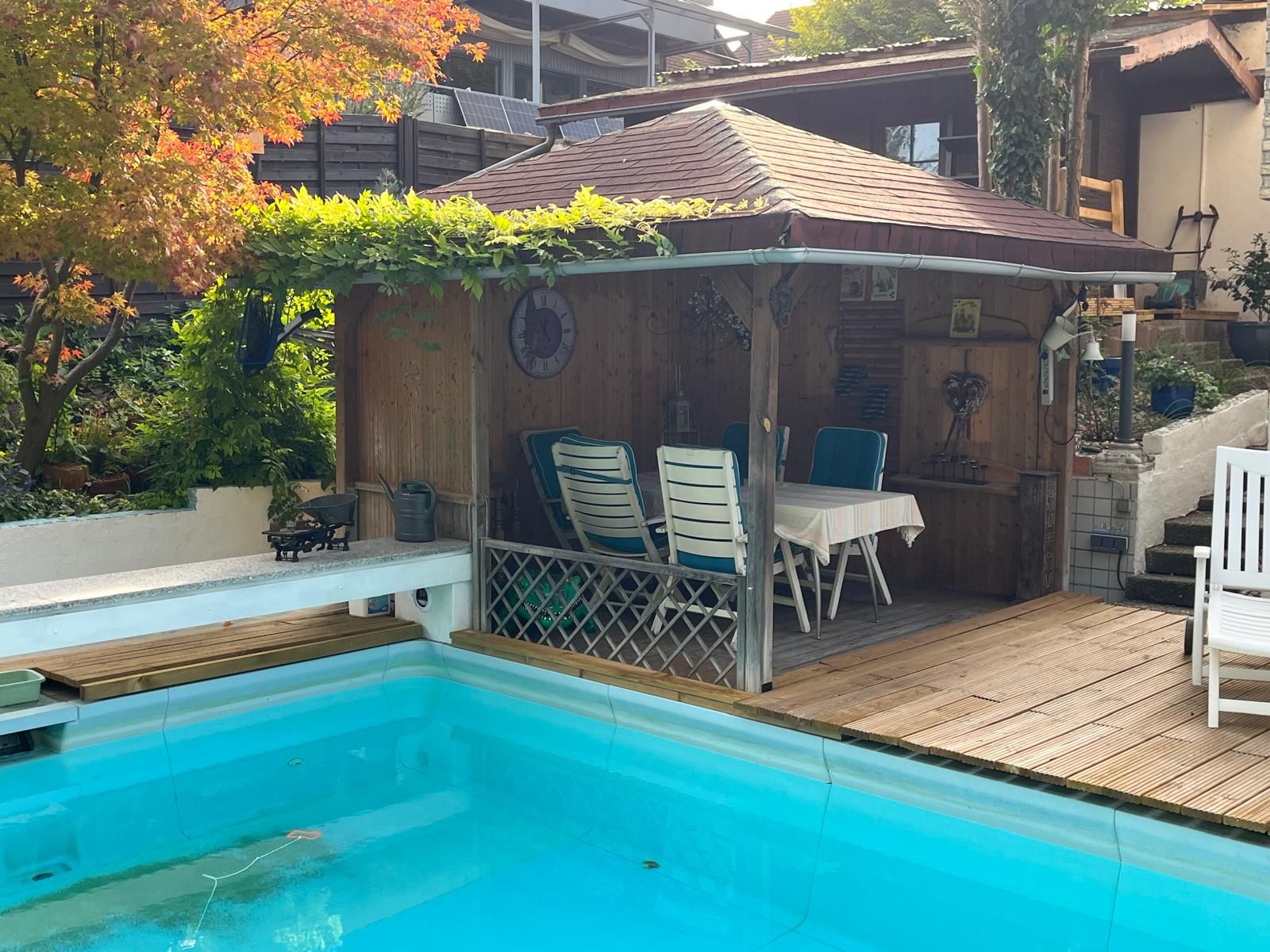 En swimmingpool med en træterrasse og et overdækket siddeområde med stole og et bord.
