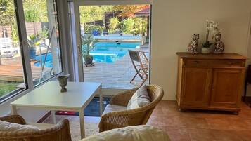 A room with a view of a swimming pool, wicker furniture, a wooden cabinet, and a small table.