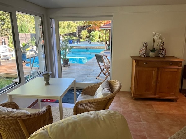 A room with a view of a swimming pool, wicker furniture, a wooden cabinet, and a small table.