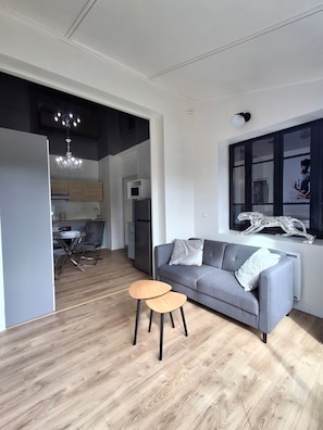 Een moderne woonkamer met een grijze bank, twee houten bijzettafeltjes, een kroonluchter en een eethoek met een tafel en stoelen.