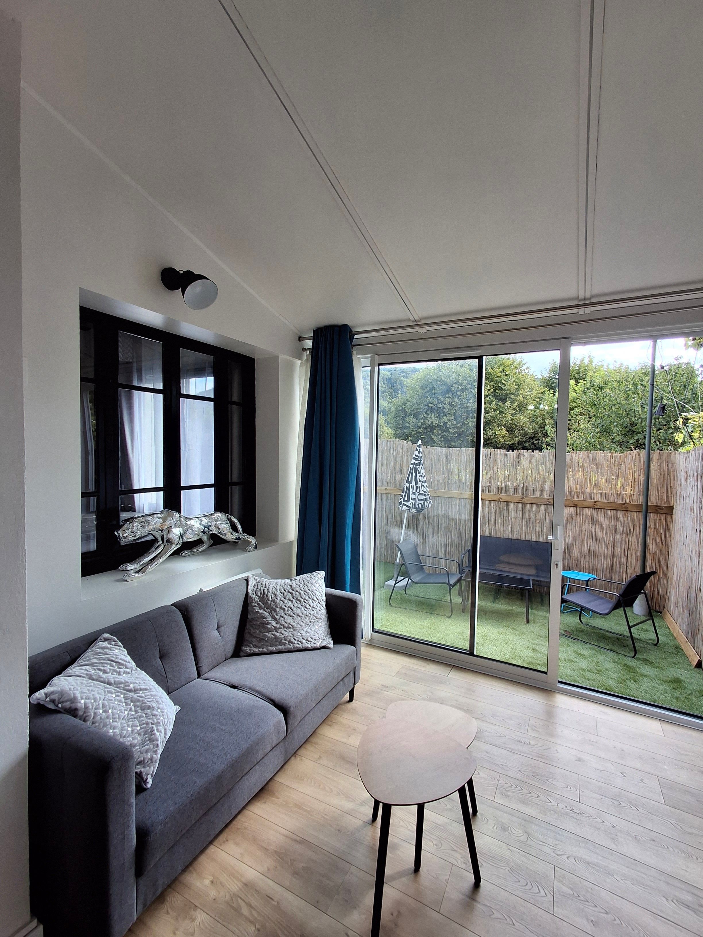 Un salon moderne avec un canapé gris, une petite table ronde et une grande porte-fenêtre coulissante donnant sur un jardin.