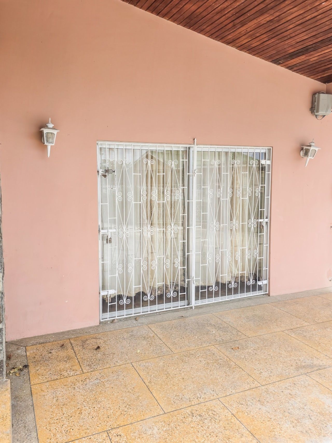 A building facade with a pink wall, a metal gate with decorative bars, and two wall-mounted light fixtures.