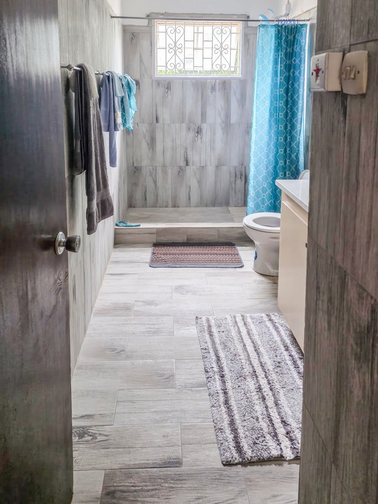 A bathroom with a shower, toilet, and a sink. There are towels hanging on a rack, a window with bars, and a patterned curtain.
