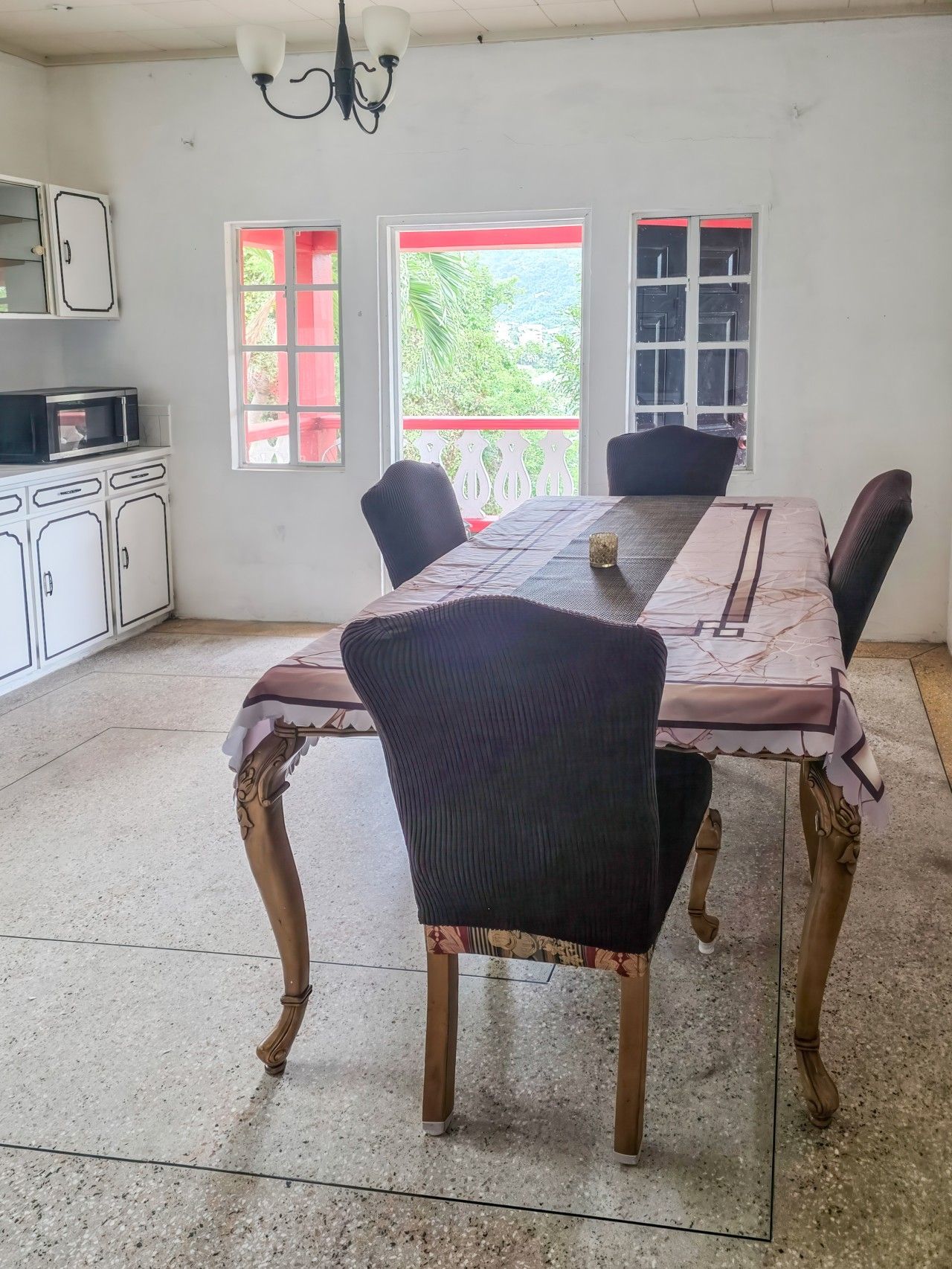 A dining area with a long table, four chairs, a chandelier, and a view of greenery outside.