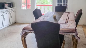 A dining area with a long table, four chairs, a chandelier, and a view of greenery outside.