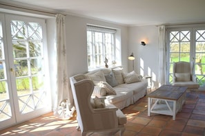 Quarto com sofá, poltronas, mesa de centro e abajur. Janelas amplas com cortinas.