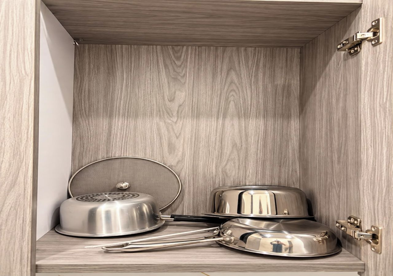 A wooden cabinet with a hinged door, containing a stainless steel pot, a pan, and a ladle.