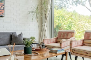 A modern living room with a white brick wall, a large window with a view of greenery, a grey sofa, patterned armchairs, a wooden coffee table with books and a decorative item, and a laptop on the left. - Staymaster Avyakta - 3BR Villa with Pool (Vagator)
