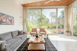 A modern living room with a sofa, chairs, a coffee table, and a large sliding glass door leading to a garden. - Staymaster Avyakta - 3BR Villa with Pool (Vagator)