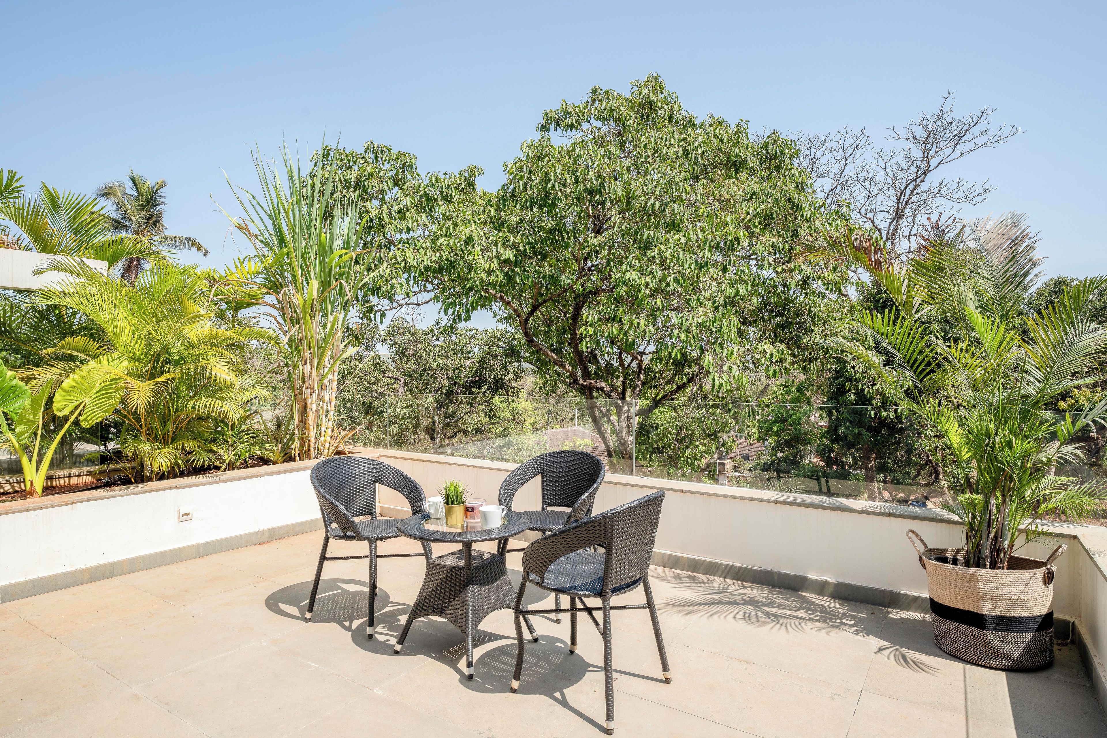 Pátio externo com mesa redonda e duas cadeiras, cercado por plantas e árvores.