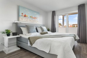 A modern bedroom with a bed, bedside tables, a painting, and a view through the window. - Holiday apartment with 2 bedrooms (Olpenitz)