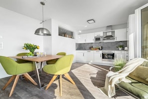 A modern kitchen with white cabinets, a microwave, and an oven. There is a dining area with a wooden table and green chairs. - Holiday apartment with 2 bedrooms (Olpenitz)