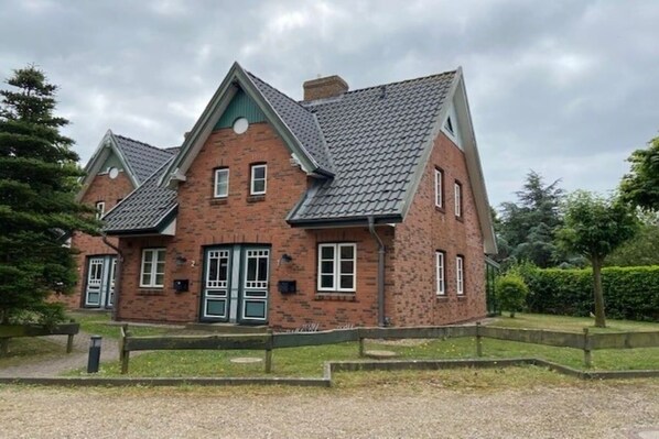 Une maison à deux étages, construite en briques, avec un toit à deux versants, une porte d’entrée verte et un petit jardin devant, délimité par une clôture en bois.