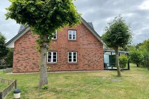 Une maison à deux étages, construite en briques, avec un toit en pente, un petit porche et un arbre dans le jardin.