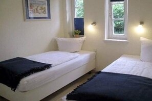 A small, simple room with two single beds, a framed picture on the wall, and a window with a view of greenery outside. - Badestraße 40a - Holiday Apartment 1 (Wyk auf Föhr)
