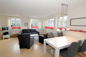 A modern living room with a sofa, a dining table, and chairs. There are windows with curtains, a radiator, and a television on a stand. - Badestraße 40a - Holiday Apartment 1 (Wyk auf Föhr)