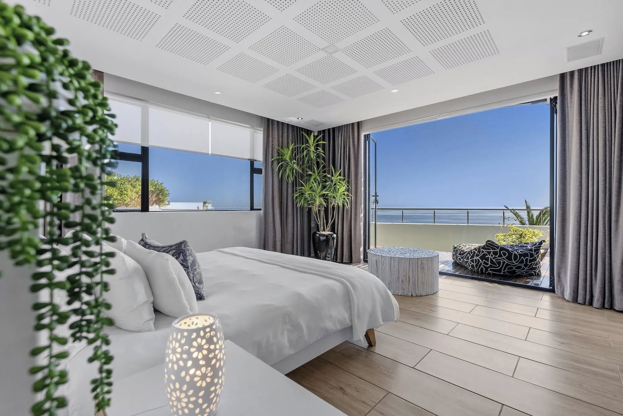 Une chambre moderne avec un grand lit, une table de chevet, une plante en pot et une vue sur la mer depuis un balcon.