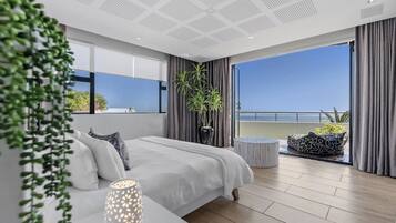 Une chambre moderne avec un grand lit, une table de chevet, une plante en pot et une vue sur la mer depuis un balcon.