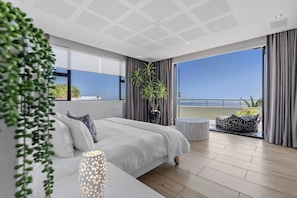 Quarto moderno com uma cama grande, uma mesinha de cabeceira, uma planta em vaso e vista para o mar através de uma varanda.