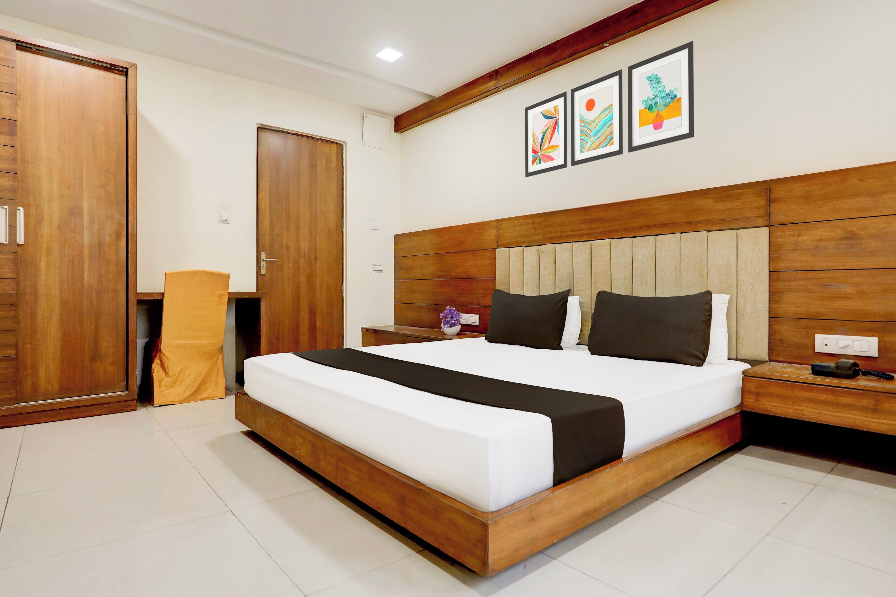 Habitación de hotel con una cama grande, cabecera de madera, una mesita de noche, un escritorio, un armario y una planta en maceta.